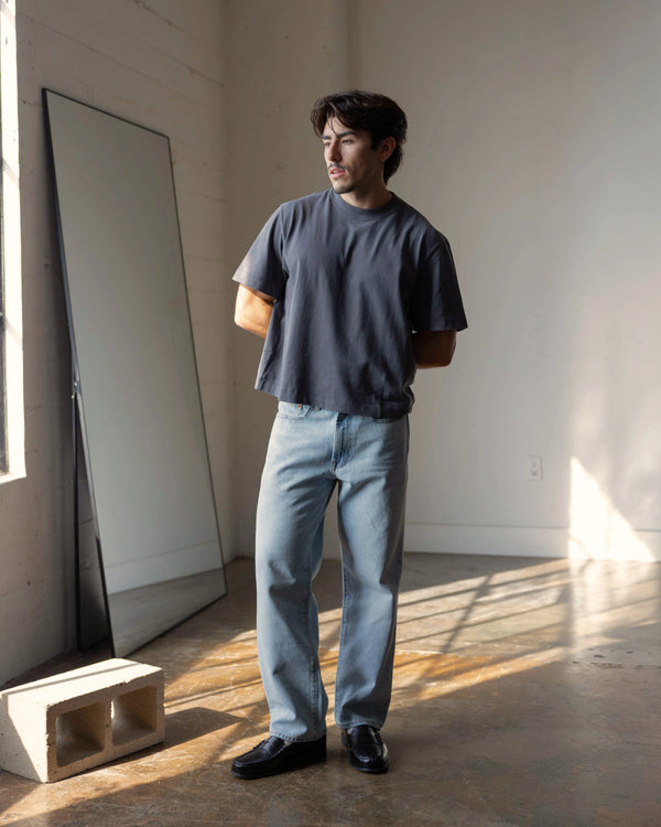 short male model wearing a dark t-shirt and light blue jeans standing in a room with sunlight streaming in.